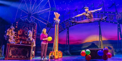 A performer leaps mid-air on a brightly lit stage with a carnival theme, while a woman holding a yellow balloon stands near a booth with stuffed animals and colorful balloons.