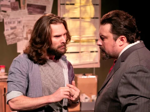 Two men facing each other in a room filled with newspapers and boxes, appearing to have a serious conversation. One man has long hair and wears casual clothes, the other has short hair and wears a suit.