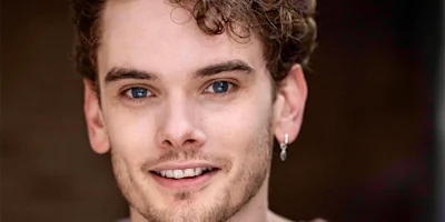 A young man with light skin, curly brown hair, blue eyes, and light facial hair, wearing a small hoop earring, smiles at the camera against a blurred background.