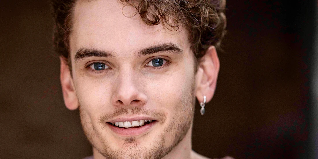 A young man with light skin, curly brown hair, blue eyes, and light facial hair, wearing a small hoop earring, smiles at the camera against a blurred background.