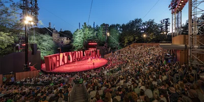 A large audience watches an outdoor theater performance at sunset, with trees and stage lights surrounding the open-air venue.