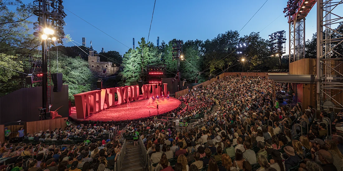 A large audience watches an outdoor theater performance at sunset, with trees and stage lights surrounding the open-air venue.