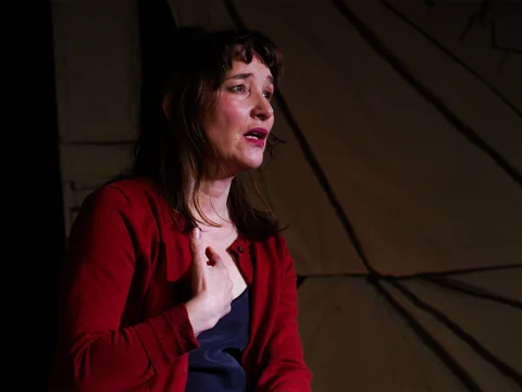 A woman with shoulder-length brown hair and wearing a red cardigan gestures with her hand on her chest, appearing to express emotion against a dark, abstract backdrop.