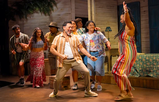 A group of people dance energetically together on a stage decorated to look like a tropical, wooden cabin interior.