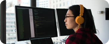 Developer working on multiple monitors displaying code. They are wearing orange headphones.