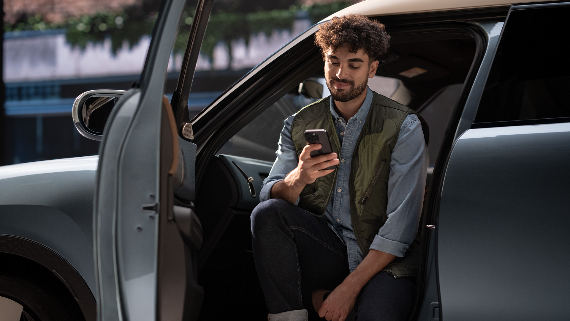 Person sitting in the driver's seat of a car with the door open, wearing a green vest and blue shirt, looking at a smartphone, with greenery and a building in the background.