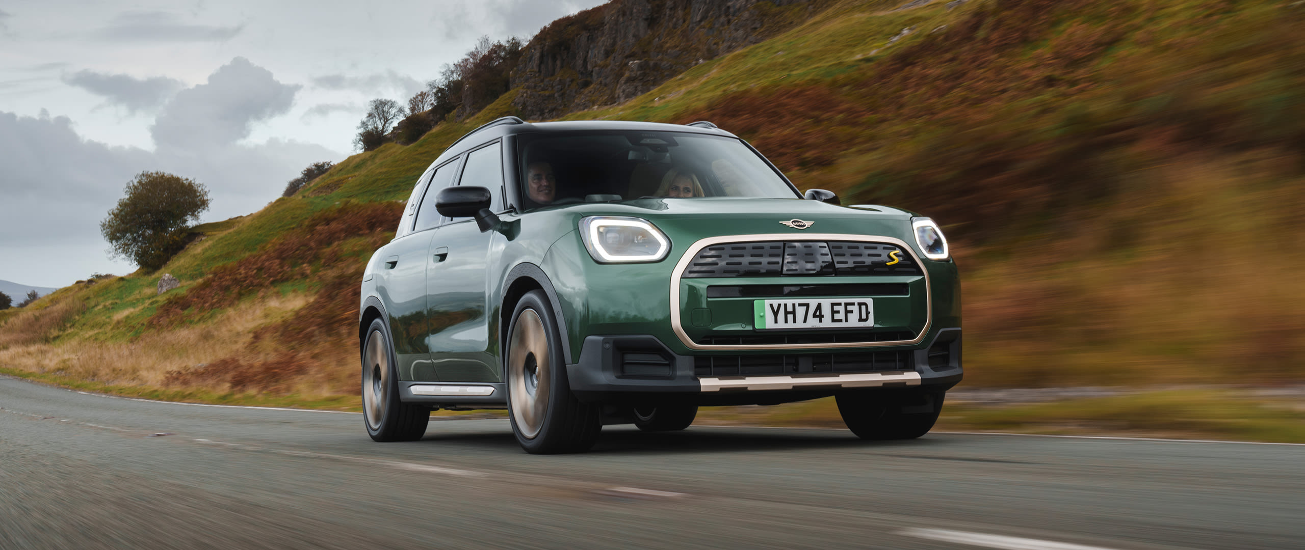 Green MINI Countryman Electric driving on a street, viewed from the front with a hill in the background.