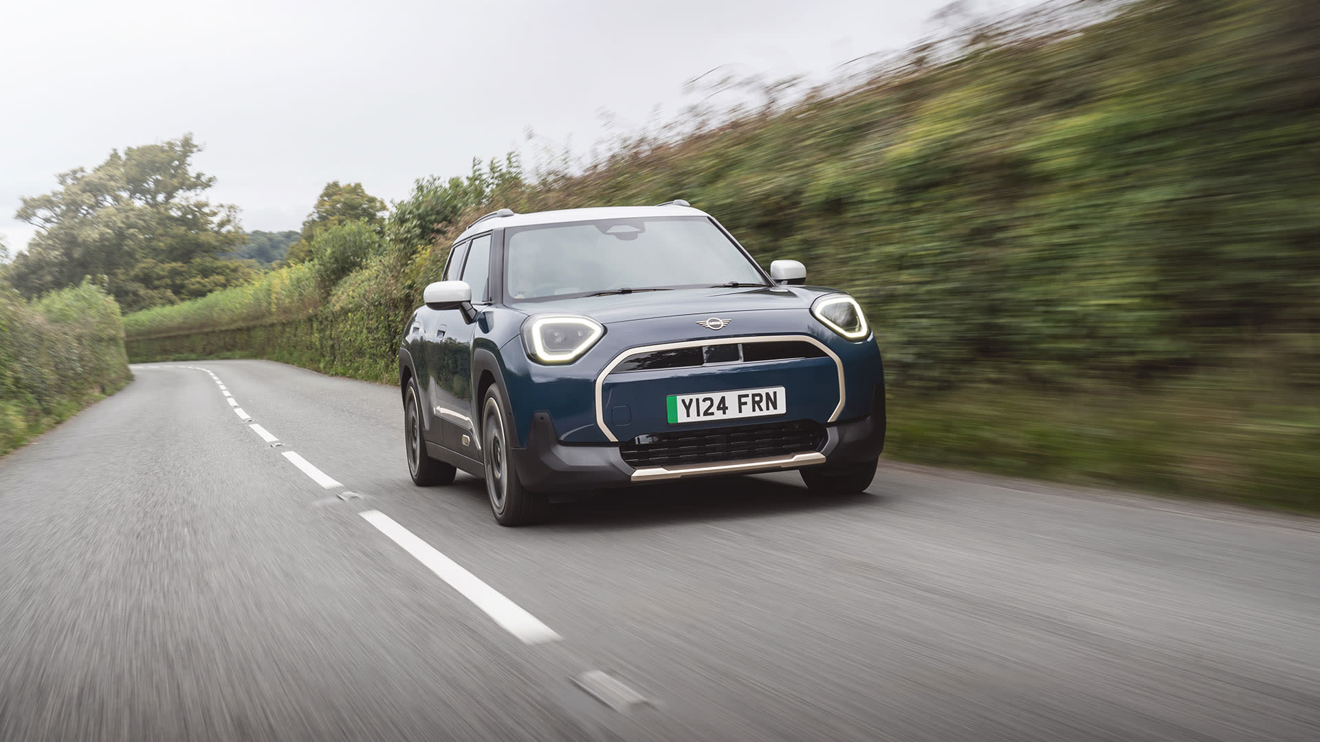 Blue MINI Countryman driving along a tree‑lined rural road.