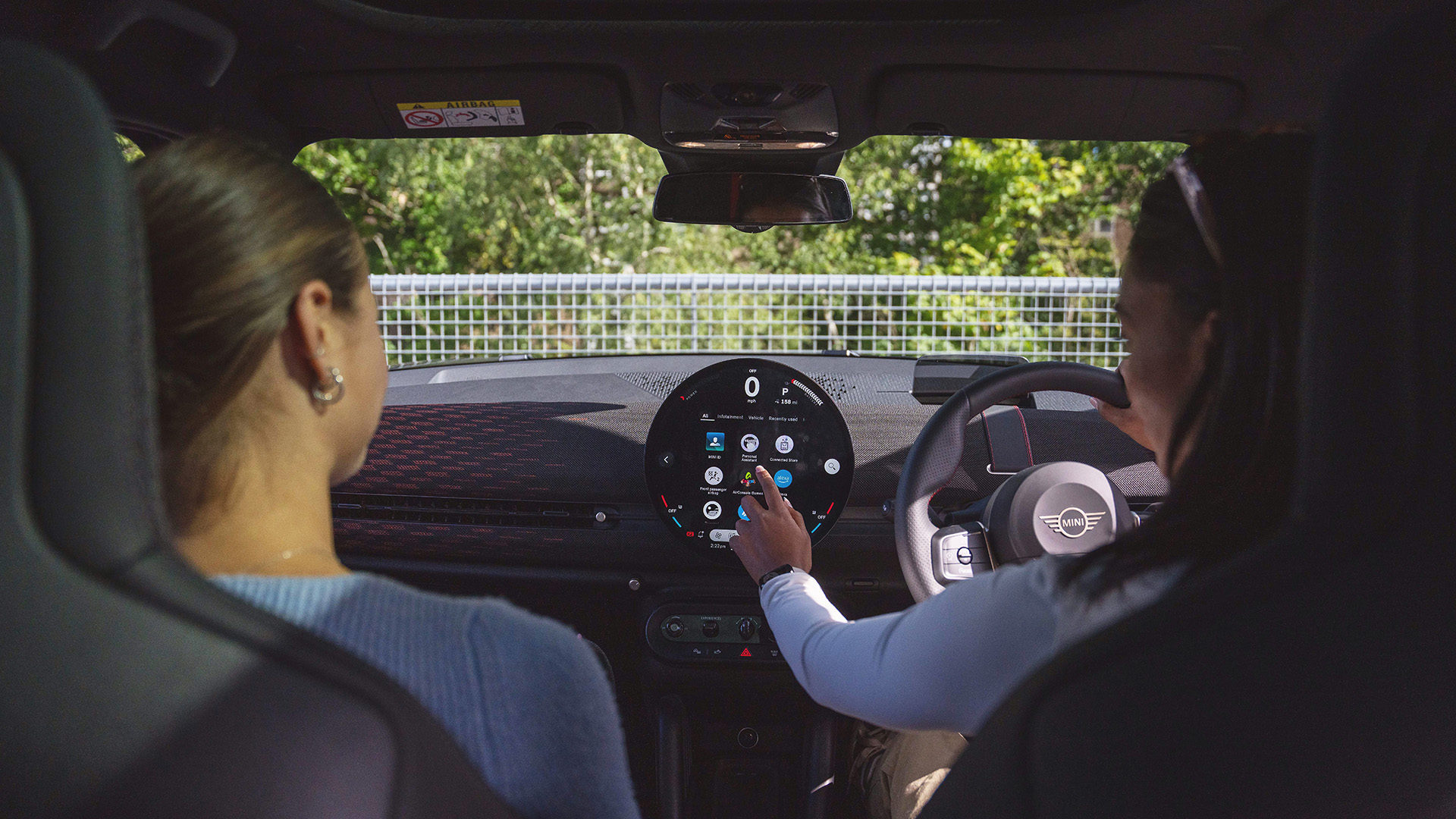 Interiour view of a MINI with two girls in front seats, and the driver making updates on the round digital interface