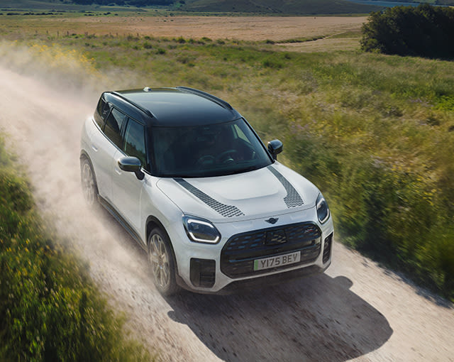 White MINI Countryman driving on a dusty countryside track, surrounded by green fields and open rural landscape.