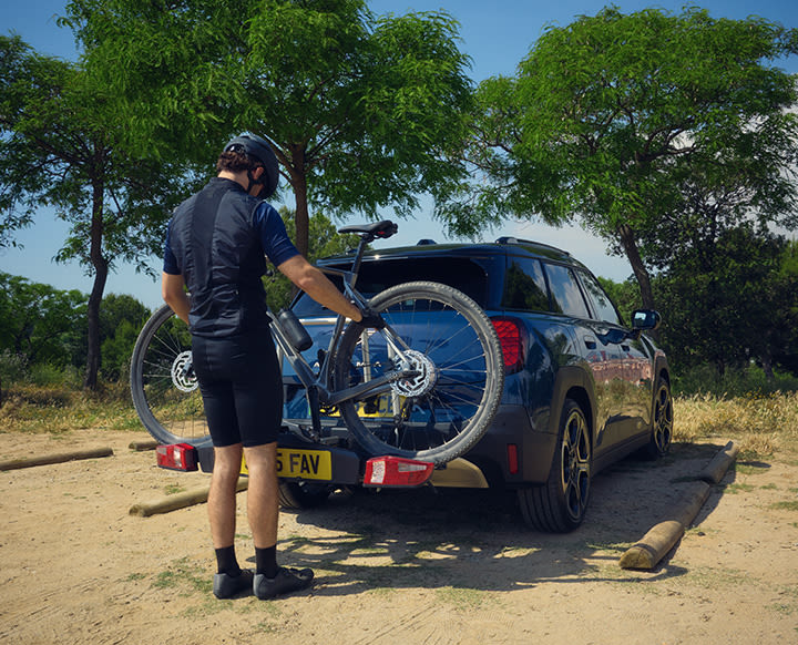 Person standing beside a MINI with a rear-mounted bike rack, securing a bicycle on a dirt clearing with trees in the background.