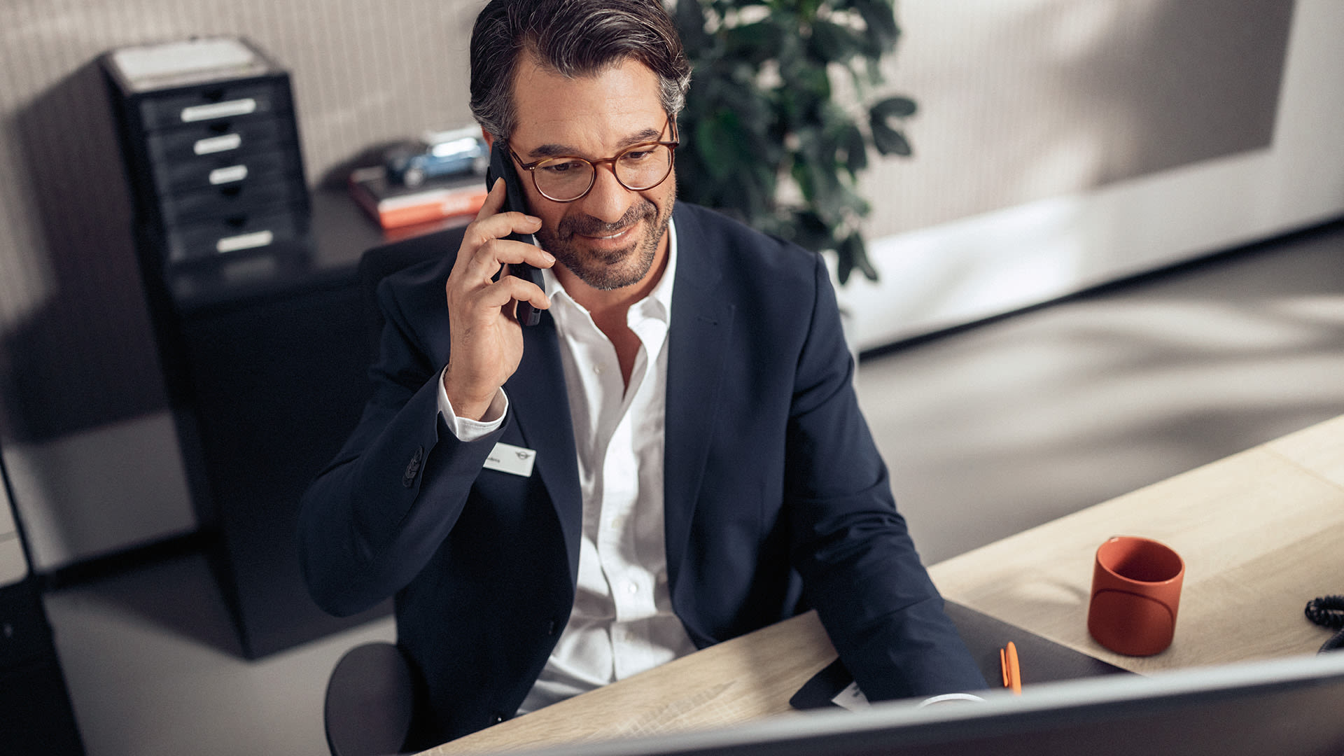 Person in business attire speaking on a mobile phone while seated at a desk in a modern office.