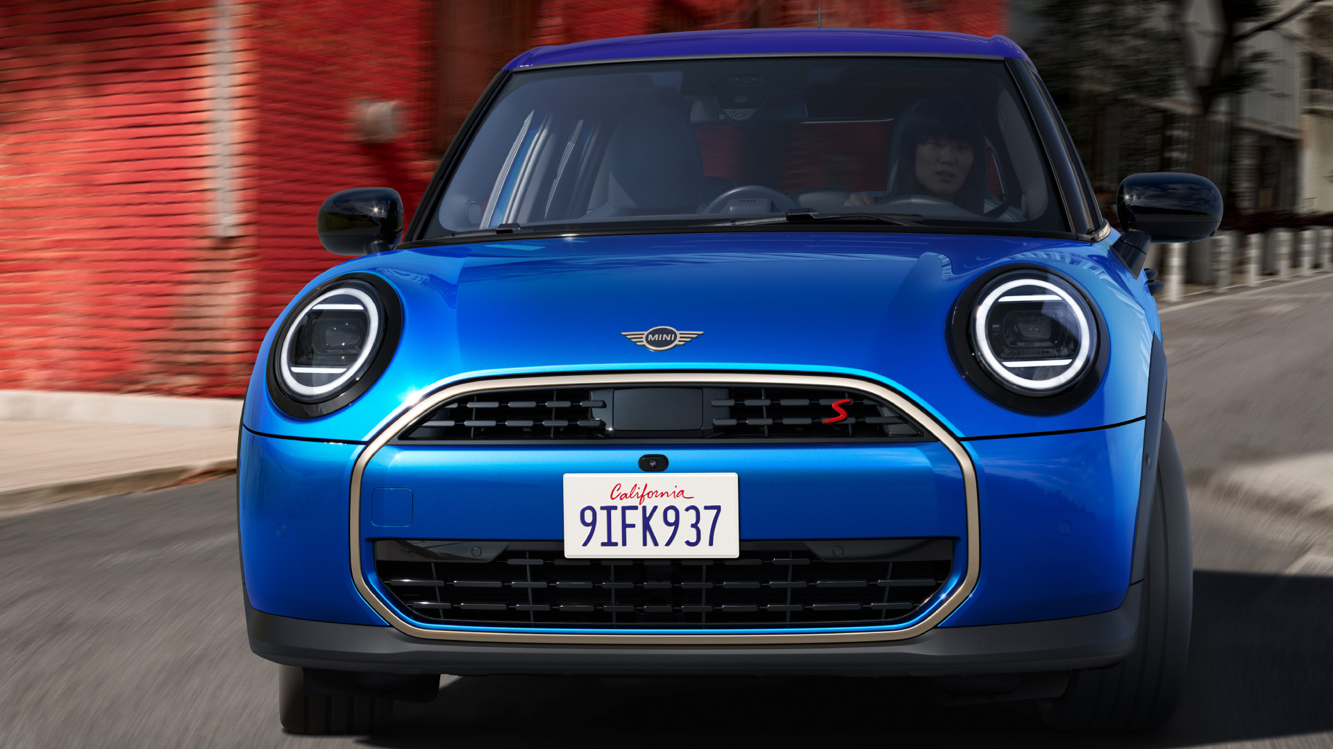 Front view of a blue MINI Cooper 5-door driving on a street with red brick buildings in the background.