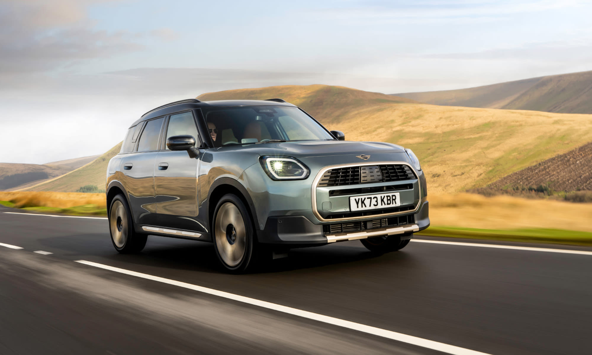 Grey MINI Countryman driving on an open road through a countryside landscape.