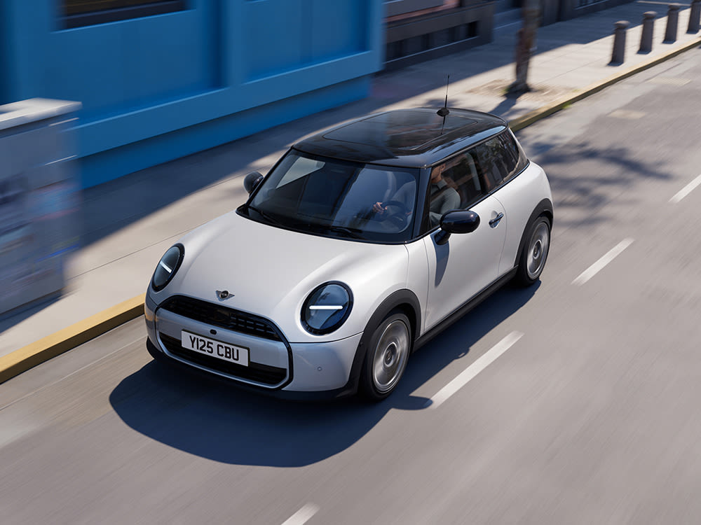 Aerial view of a light gray MINI Cooper driving on a city street next to a blue wall building