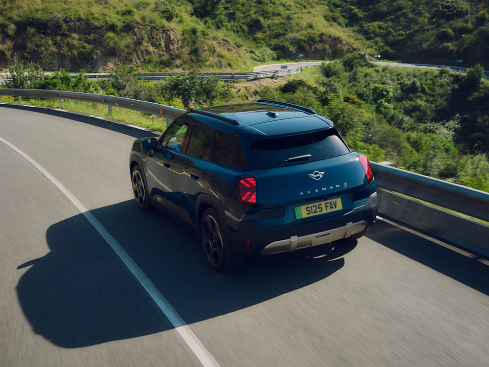 A blue MINI Aceman S driving on a winding mountain road with lush green hills in the background.
