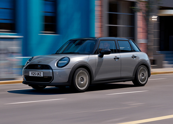 Left side view of a gray MINI Cooper 5-door driving on a street with buildings in the background.