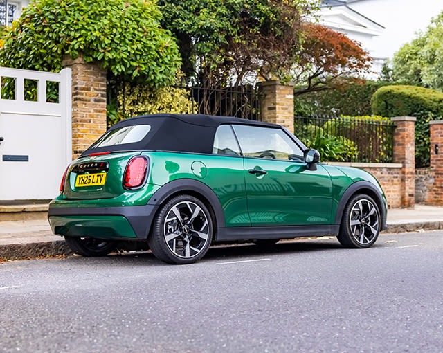Green MINI Convertible with black soft top parked on a residential street beside brick walls and garden hedges.