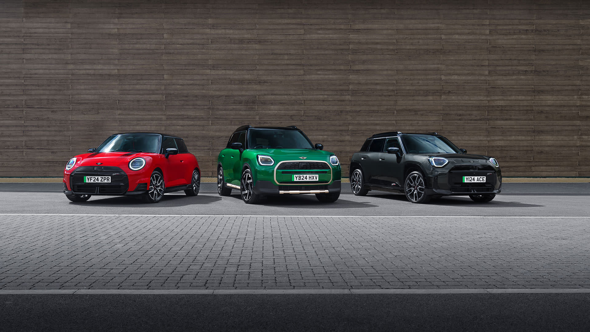 Three MINI cars parked side by side in front of a pink wall. From left to right, the cars are blue, orange, and silver.