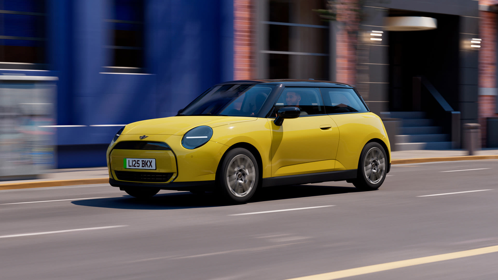 Left Front-side view of a Yellow MINI Cooper electric driving on a street with blue building in the background