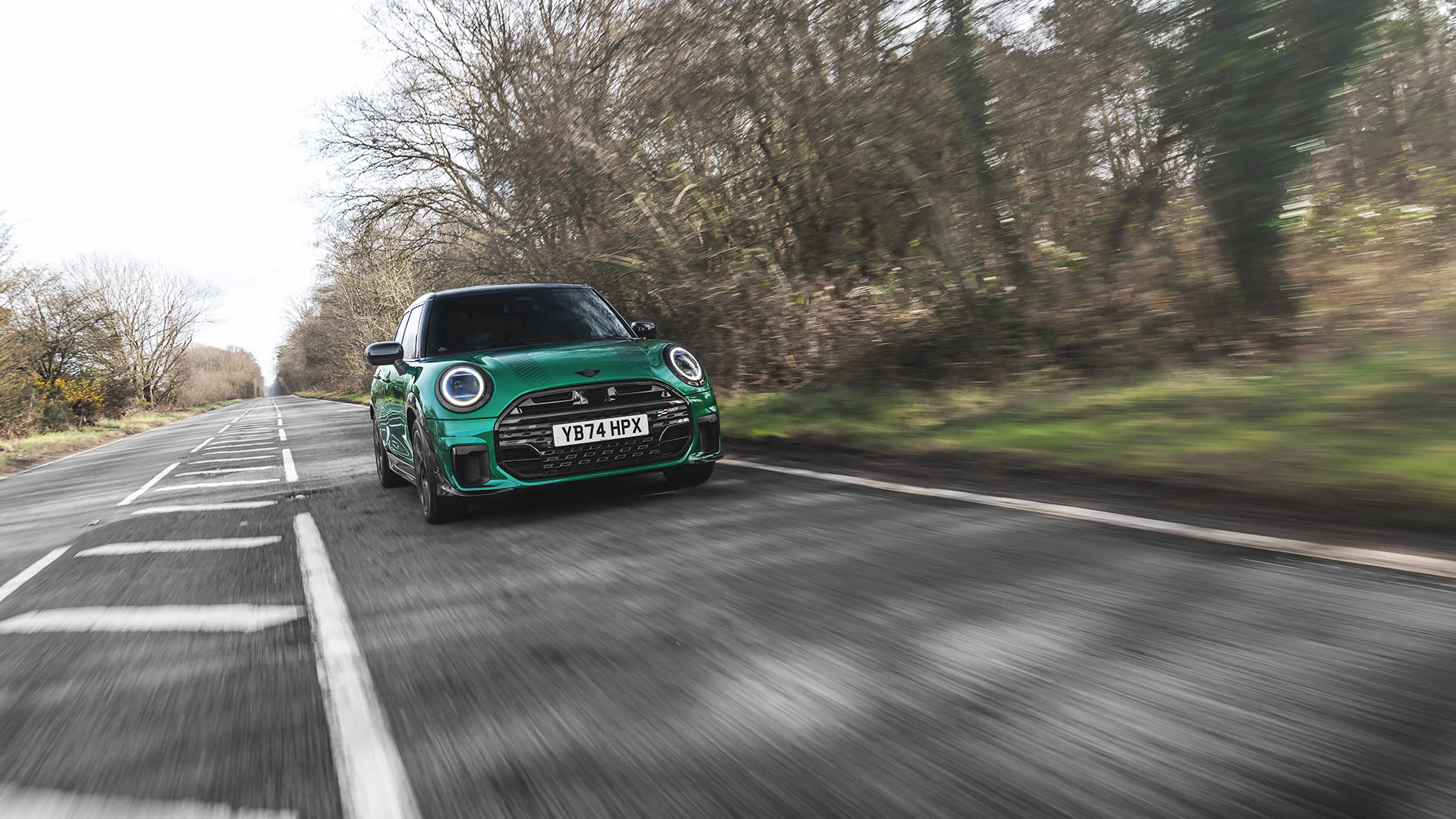 Green Mini Cooper hatchback driving along a tree-lined rural road, captured from a low front angle with motion blur in the background and road markings emphasising speed.