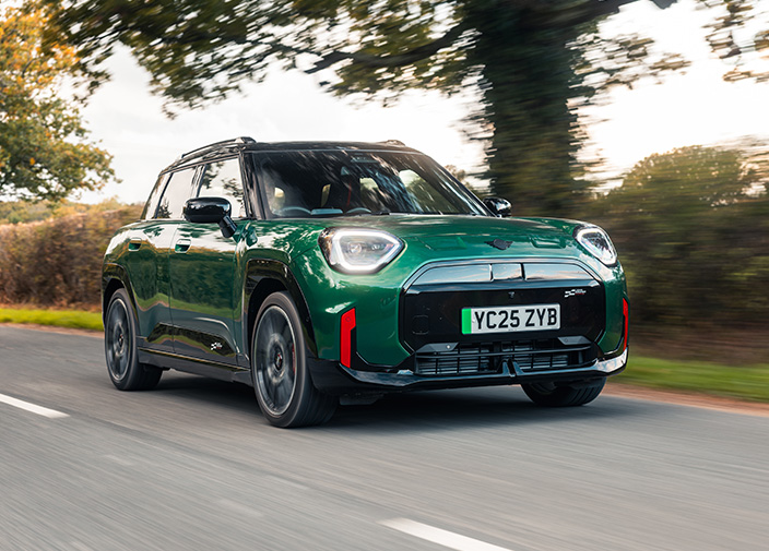 Front view of a green MINI Aceman on a road with hedge and trees in the background