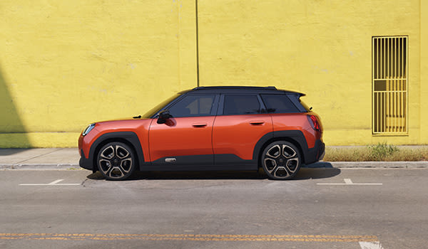 Orange MINI Aceman parked on a street in front of a bright yellow building with a tiled roof and a small shop entrance.