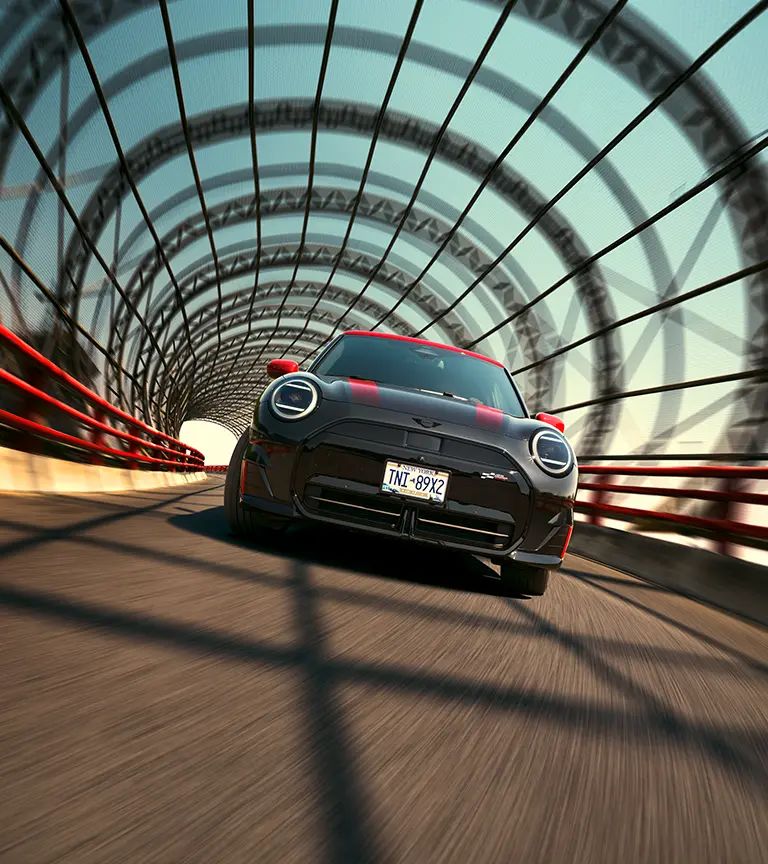 A black MINI Cooper with red racing stripes driving through a tunnel with a geometric lattice structure and red railings.