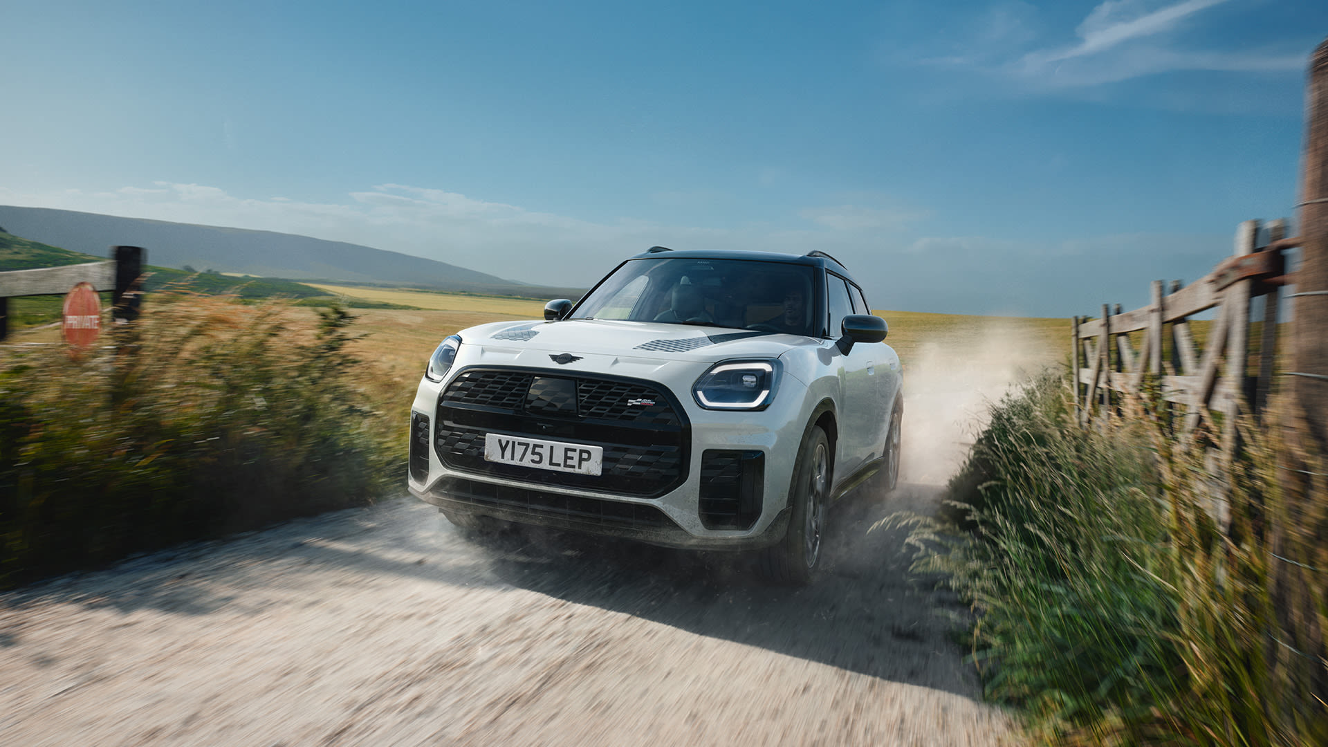White Mini Countryman SUV driving quickly along a narrow country lane, kicking up dust beside wooden fences and grassy fields, with open countryside and blue sky in the background.