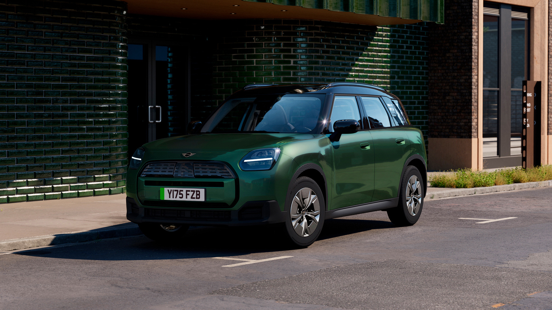 Green MINI Countryman electric parked on a city street in front of a modern building with green tiles and large windows.