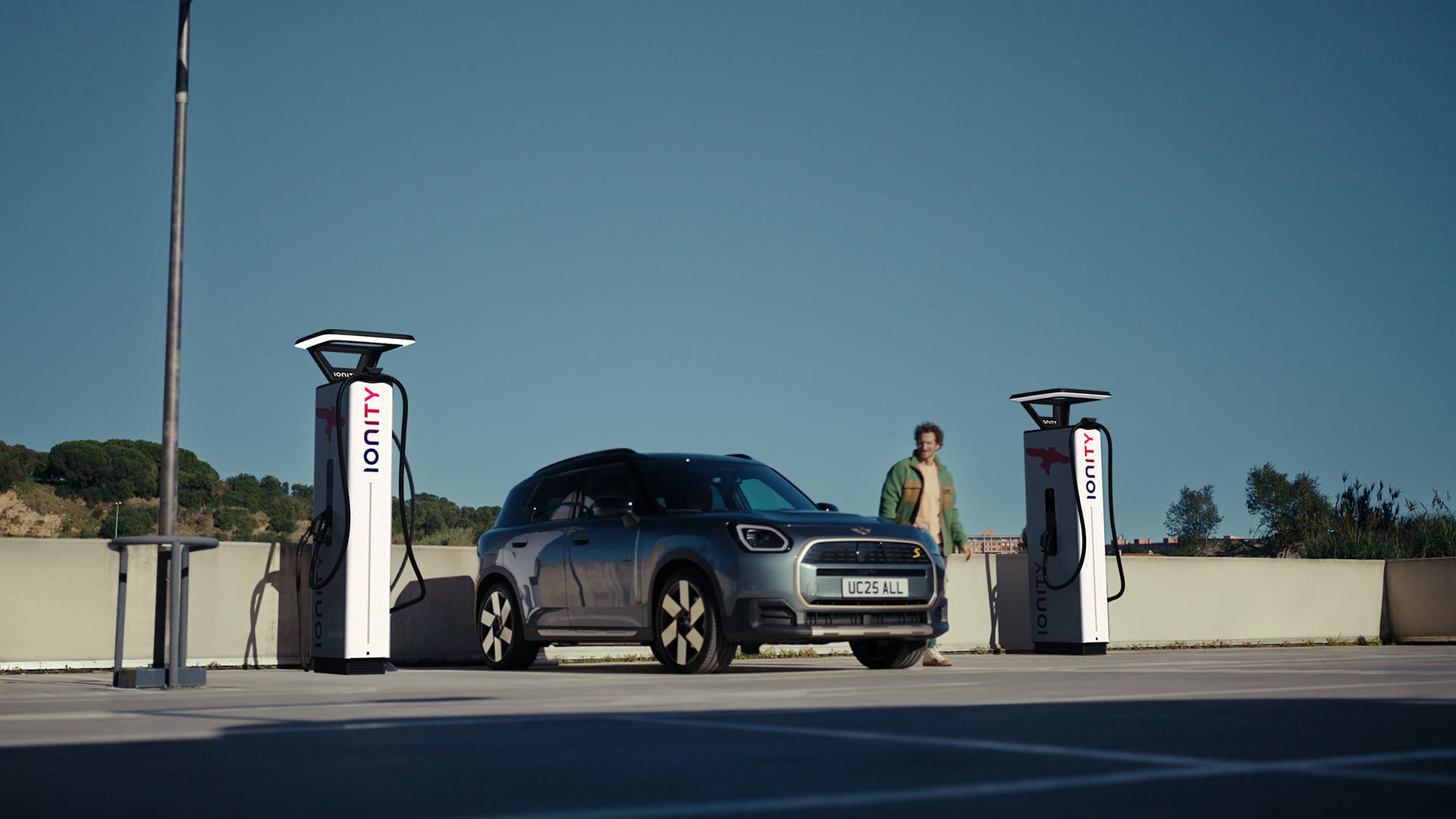 MINI Aceman electric crossover charging between two Ionity stations in an outdoor car park under a clear blue sky.