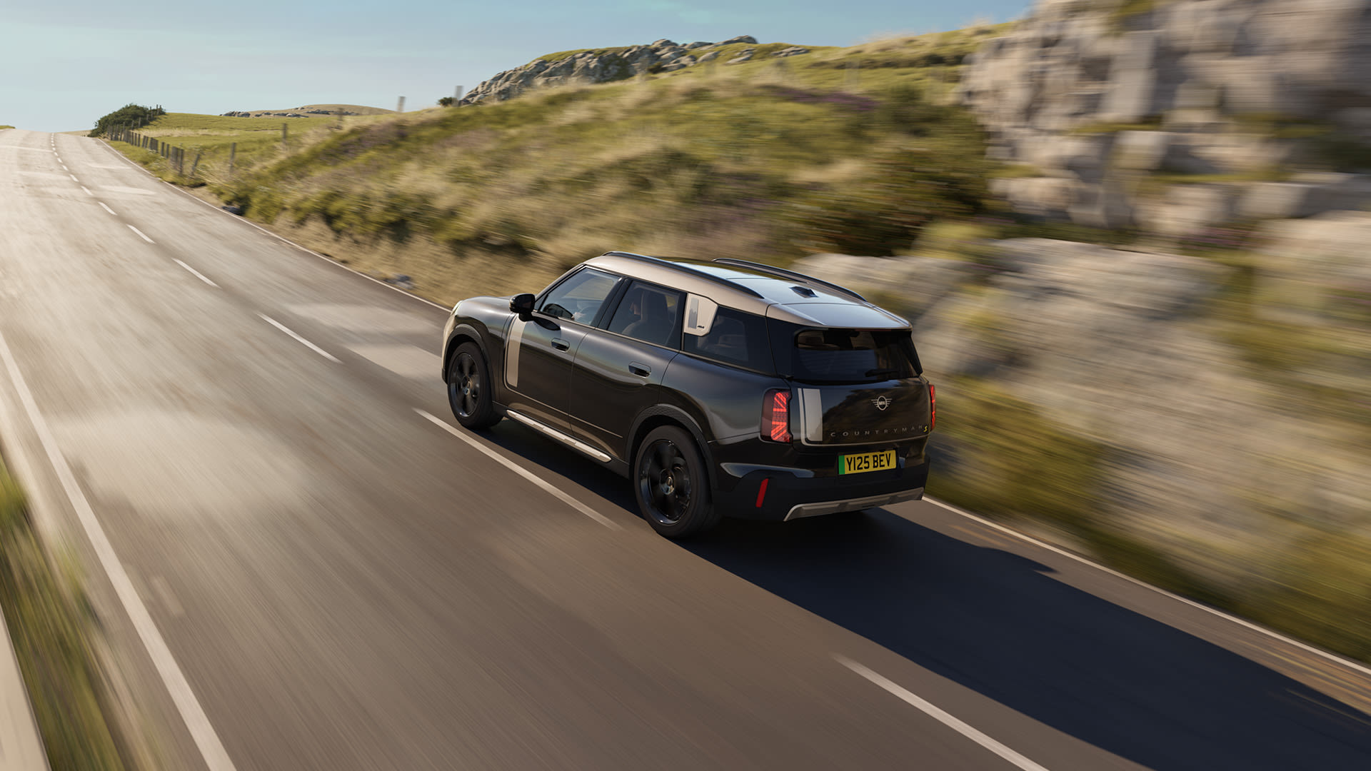 Black Mini Countryman driving along a winding rural road, captured from behind at speed, with motion blur in the landscape of grassy hills, rocks, and fencing under clear daylight skies.