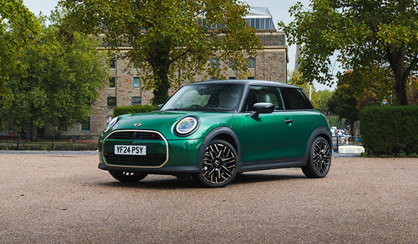 Green MINI Cooper parked on a paved area surrounded by trees, with a stone building in the background.
