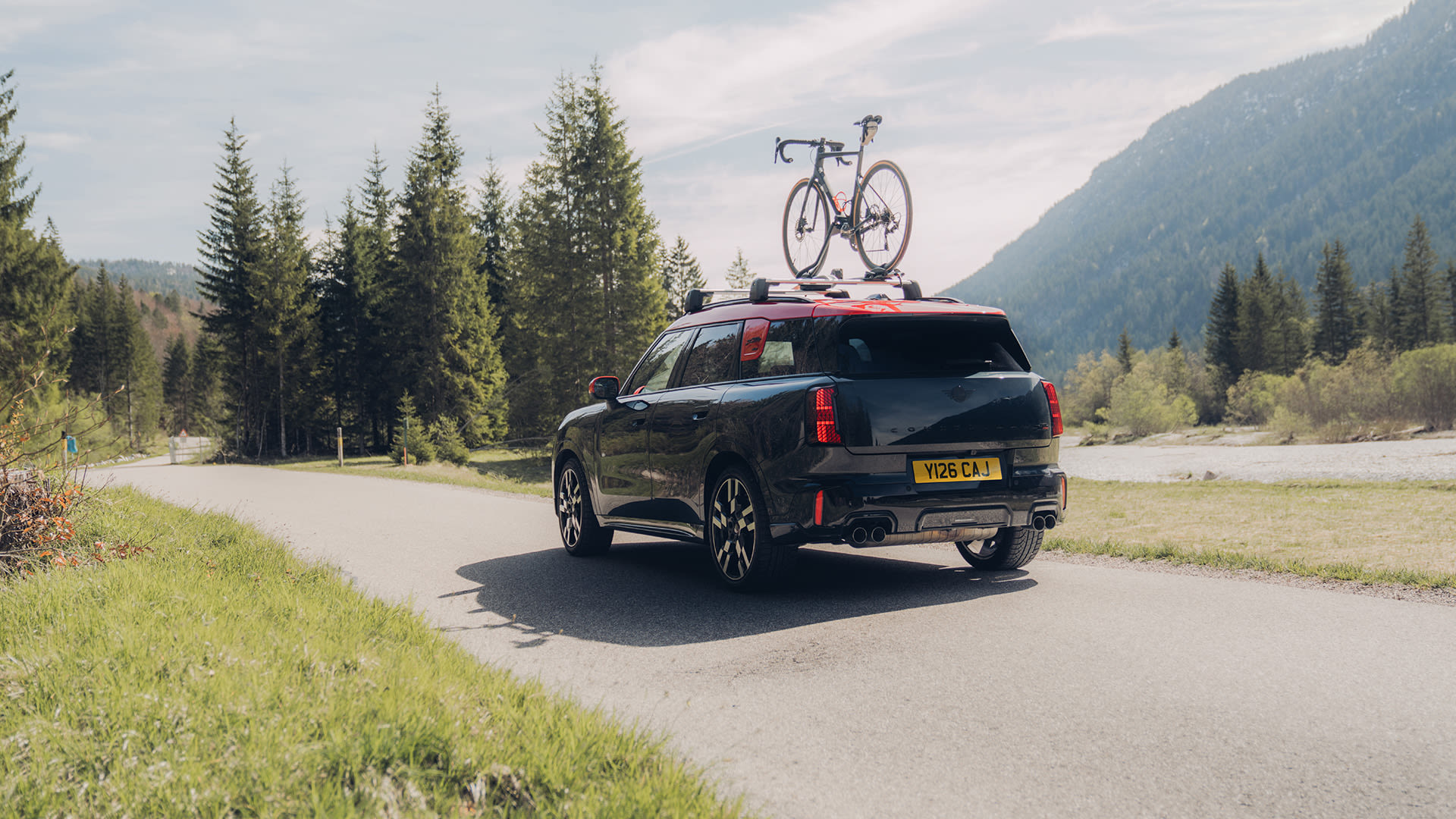 Black MINI Countryman driving on a rural road with a bicycle mounted on the roof, surrounded by trees and mountains.