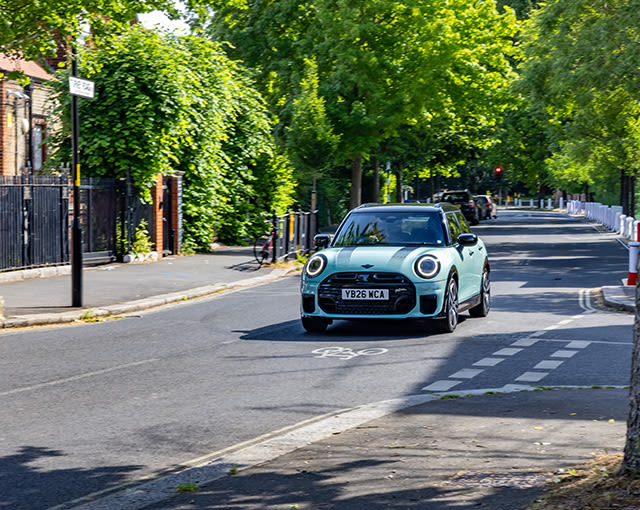 Light blue MINI Cooper driving along a quiet, tree-lined residential street on a sunny day.