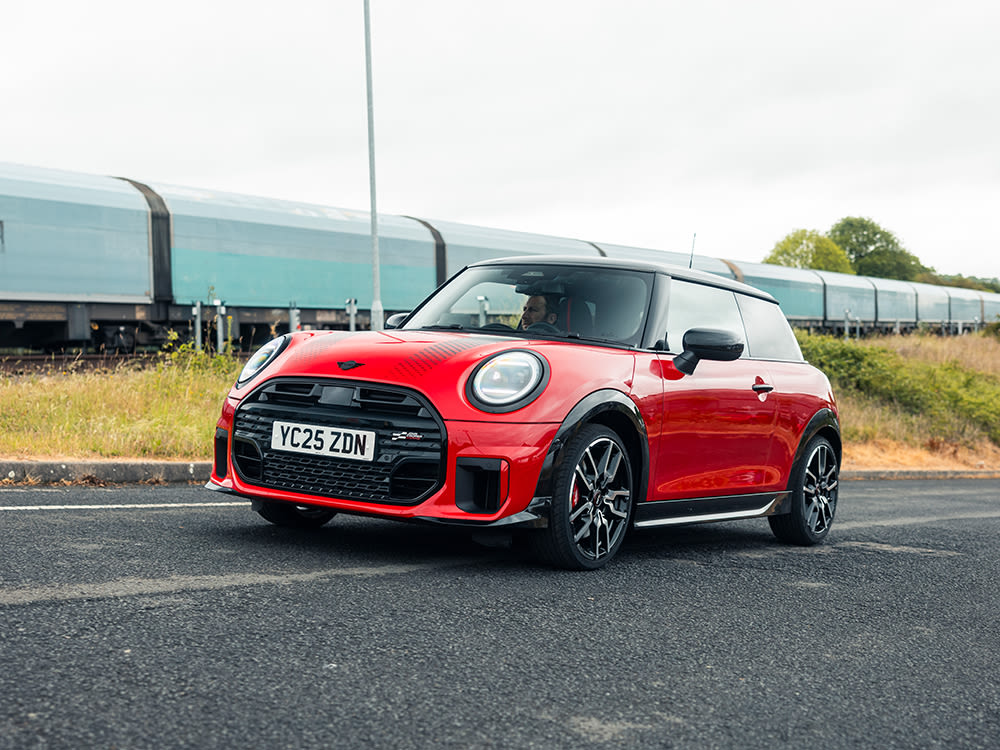A red 3-door John Cooper Works with a black roof and black wheels parked on a road, with a train passing by in the background.