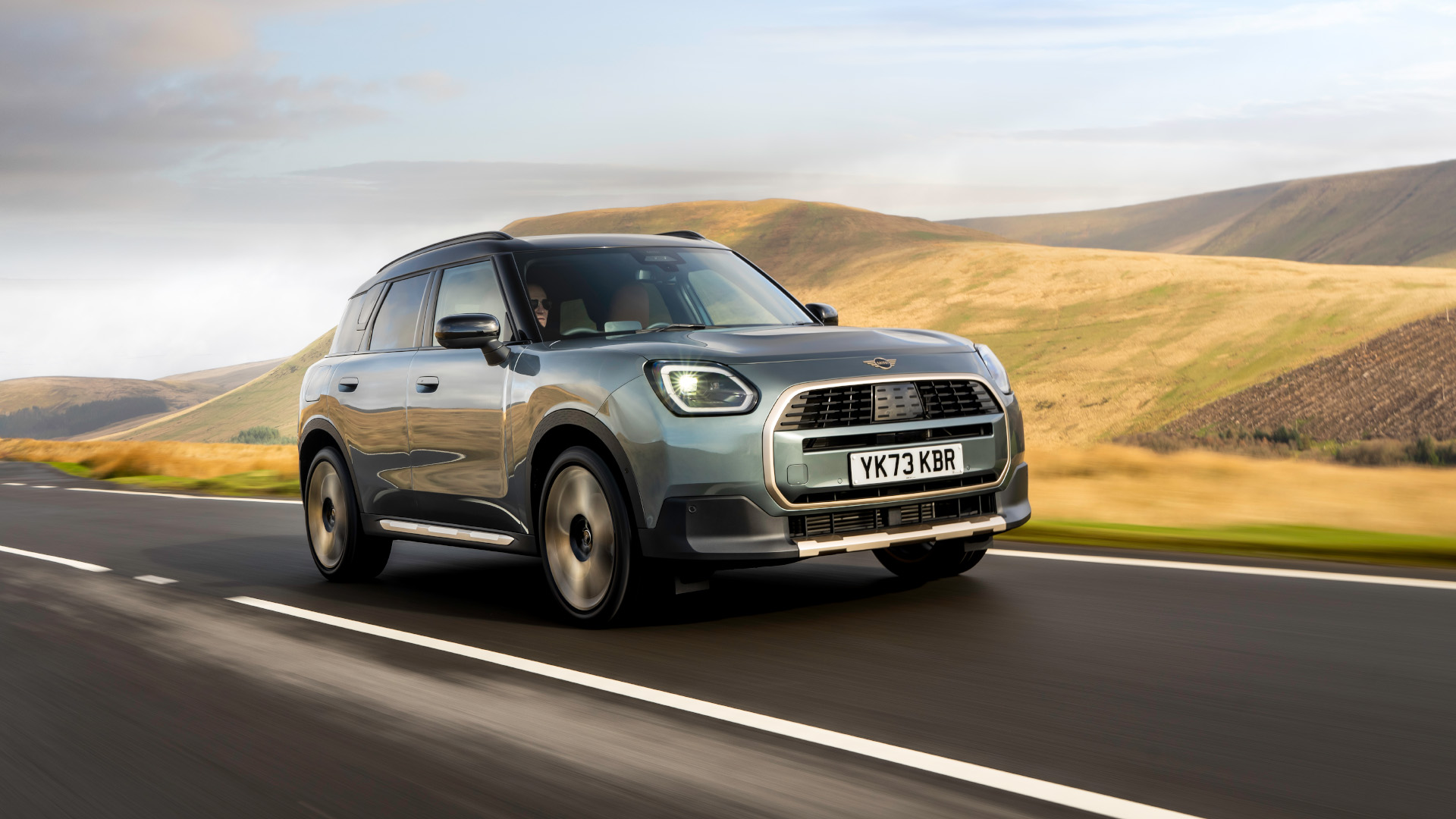 A silver MINI Countryman Electric driving on a scenic road with rolling hills and a partly cloudy sky in the background.