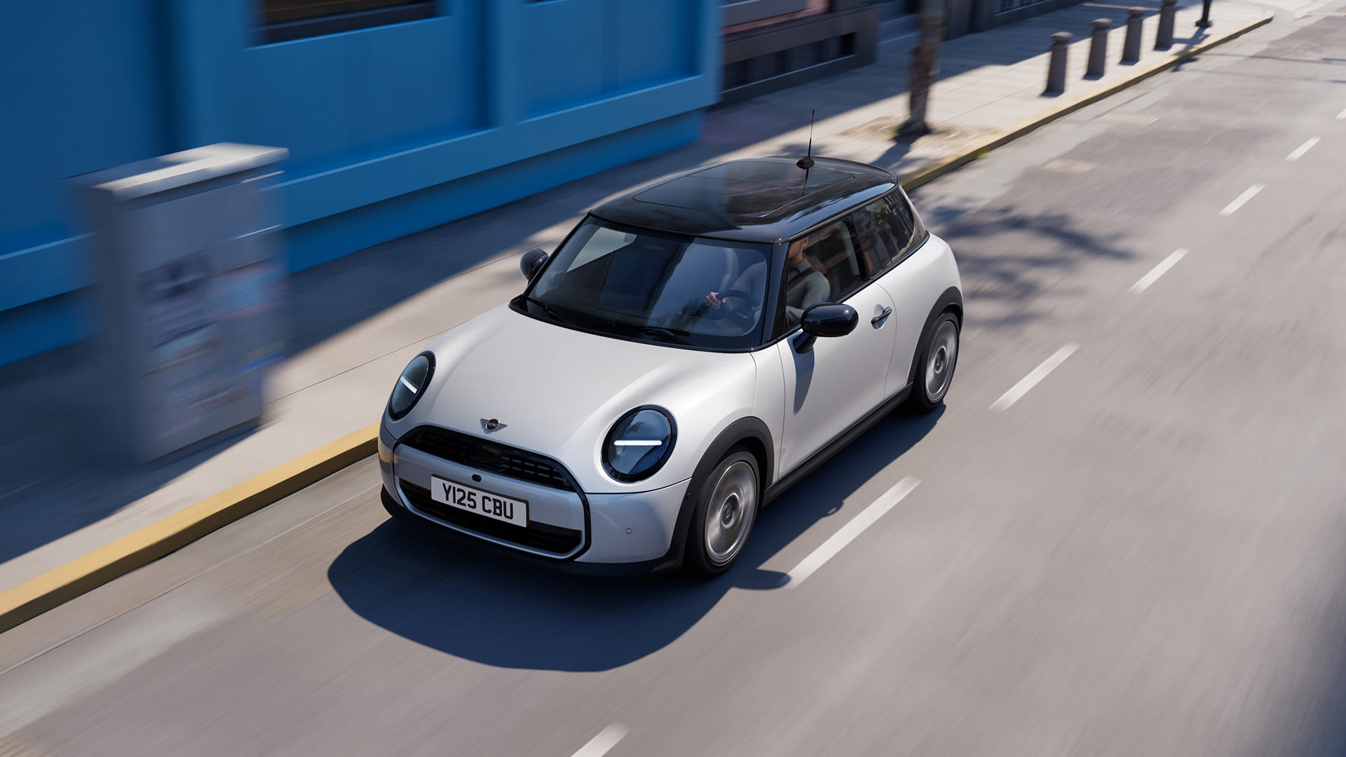 White Mini Cooper hatchback driving along a city street, viewed from above at an angle, with motion blur showing movement past a blue building and pavement bollards in bright daylight.