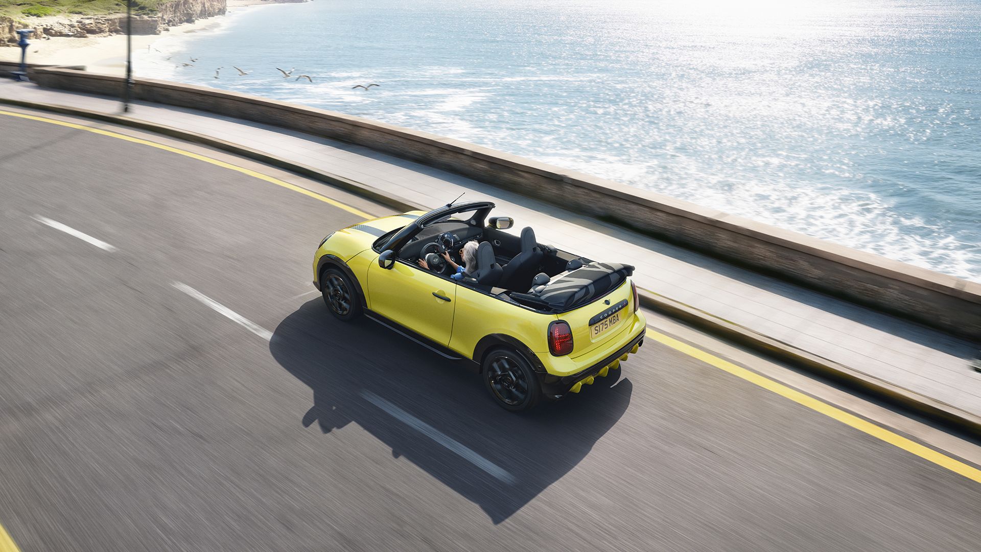 Yellow MINI Cooper Convertible driving along a coastal road with the roof down beside the sea.