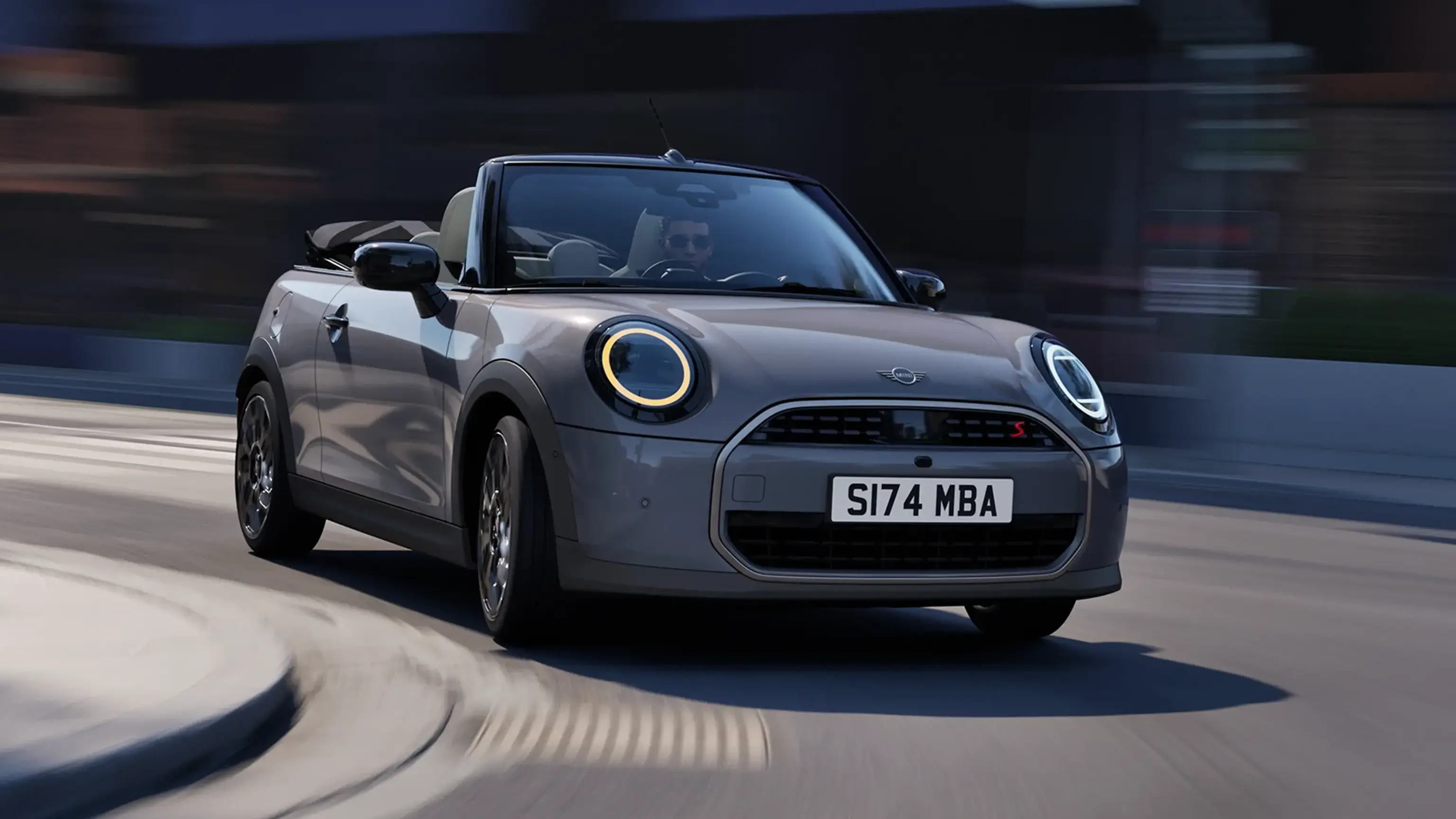 Grey MINI Cooper Convertible with the top down and headlights on, driving on a city street. 