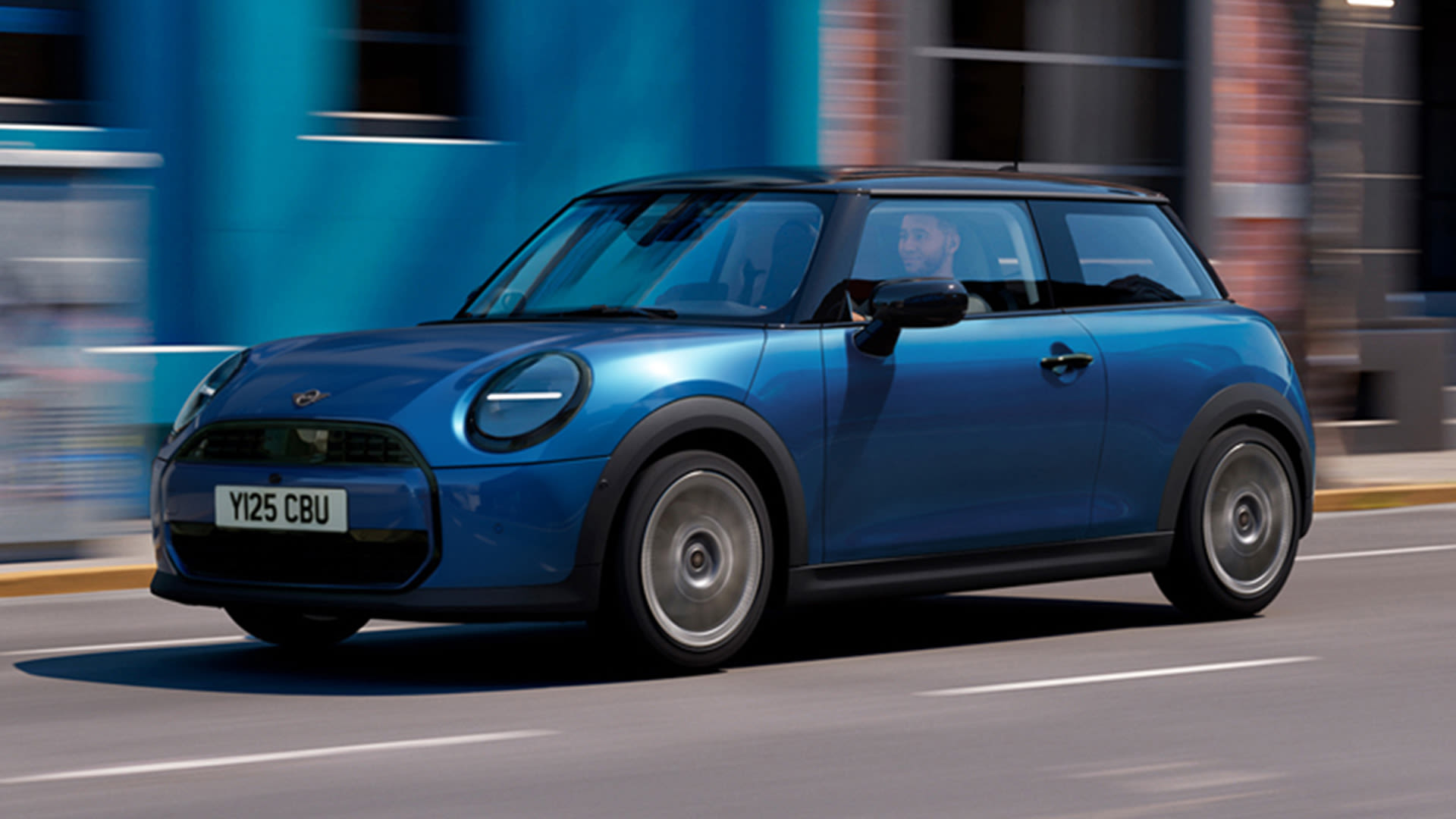 Side view of a blue MINI cooper 3 door on a street with a blue and red building in the background