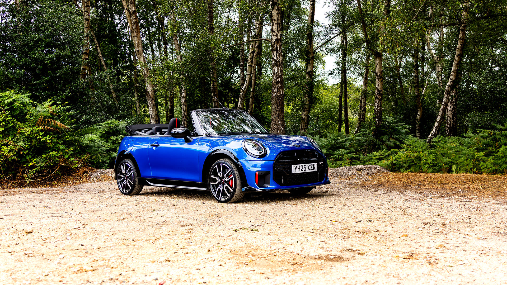 Blue Mini convertible parked on a sandy clearing in a forest, surrounded by tall trees and green foliage, with the roof down and natural daylight filtering through the canopy.