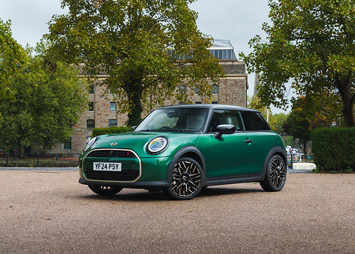 Front view of a green MINI Cooper on a gravel lot with trees and a big mansion in the background