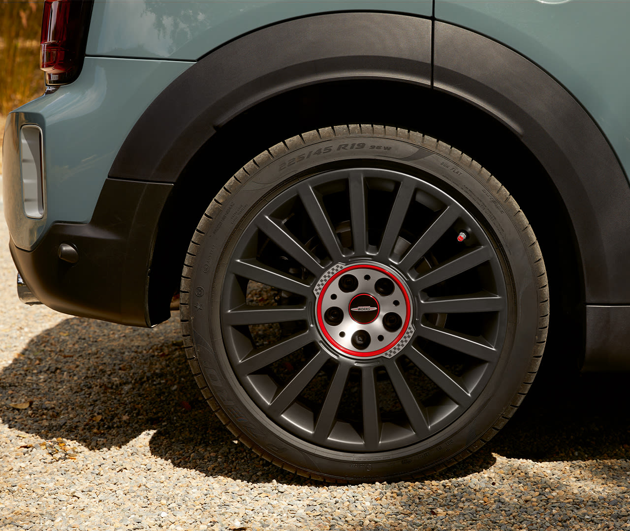 Close-up of a car's rear wheel with a black alloy rim and red accents, parked on a gravel surface.