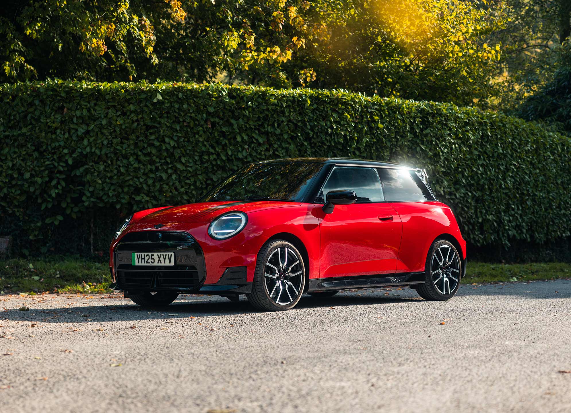 Red MINI Cooper JCW on a gravel road, in front of a green hedge with trees in the background