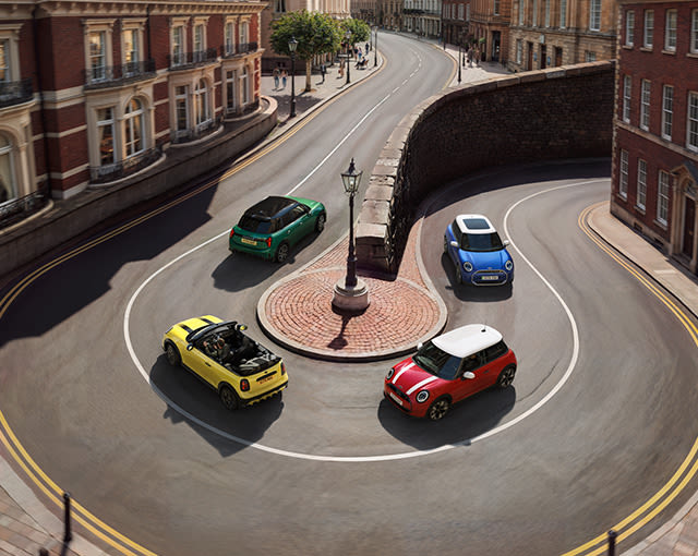Four colourful MINI cars drive around a curved city junction, surrounded by historic brick buildings and a central street lamp.