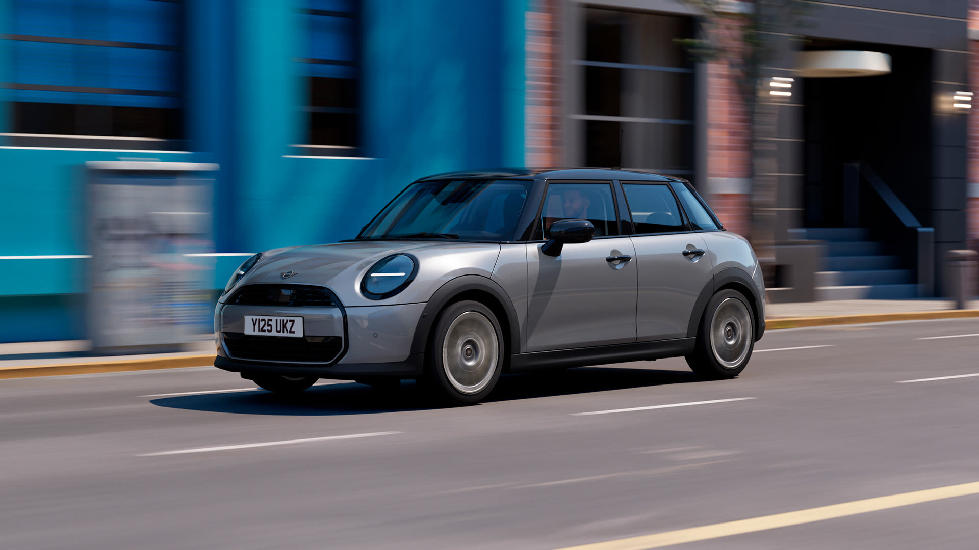 A silver MINI Cooper 5-door driving on a city street with motion-blurred buildings in the background.