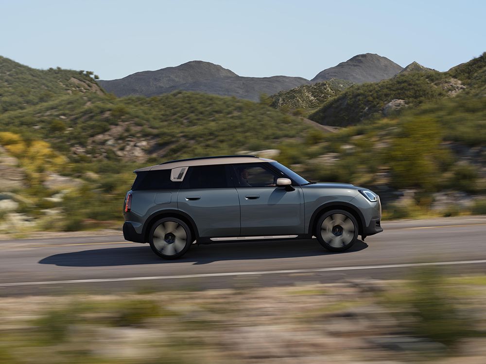 Side view of a MINI Countryman electric car driving on an open road through a hilly landscape.