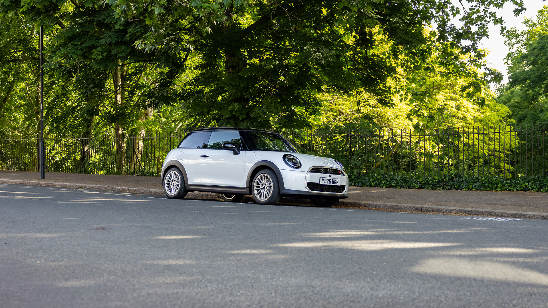 White Mini hatchback parked on a quiet roadside beside a black iron fence, with dense green trees and sunlight filtering through leaves in the background.