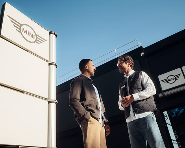 Two people talking outside a MINI dealership, with prominent MINI signage against a clear blue sky.